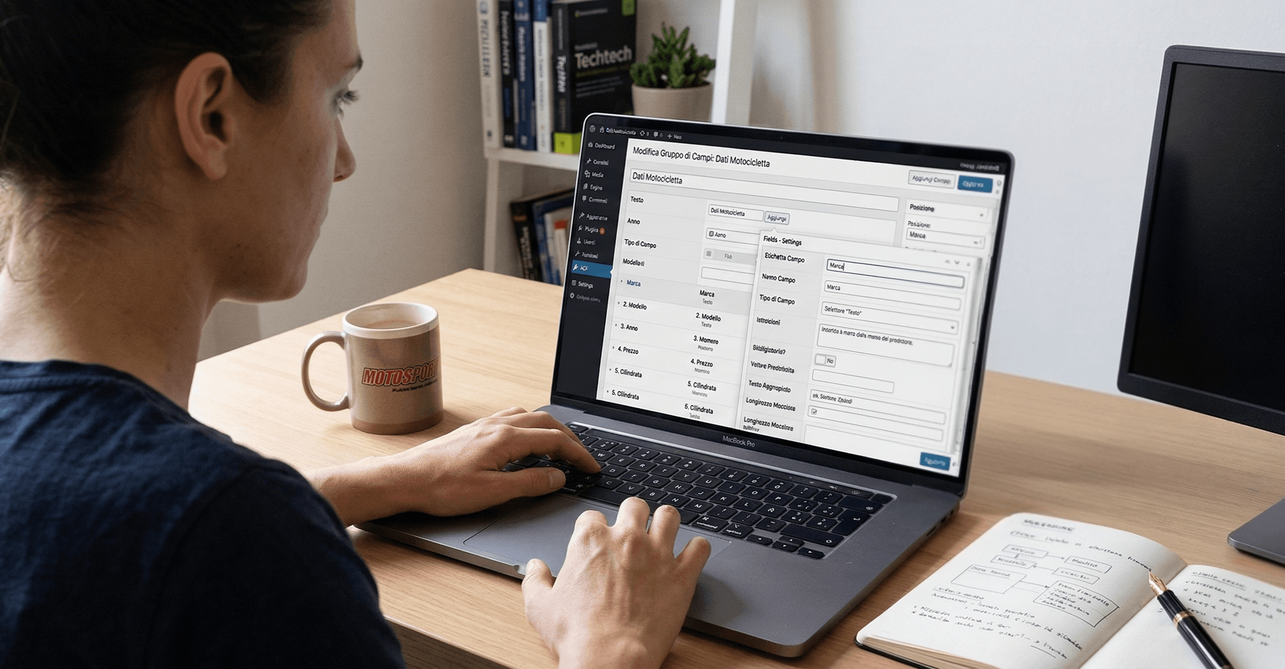 Una sviluppatrice davanti a un MacBook che mostra l'interfaccia di WordPress con ACF, su una scrivania con quadernetto, tazza e monitor esterno